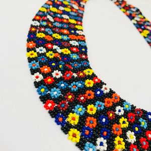 May include: Multi-strand beaded necklace with a floral pattern. The necklace is made of small, colorful beads in shades of red, orange, yellow, blue, white, and black. The beads are arranged in a repeating pattern of flowers.