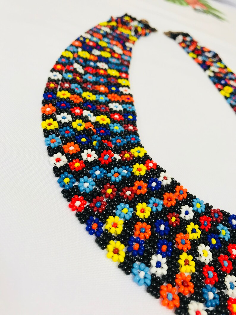May include: Multi-strand beaded necklace with a floral pattern. The necklace is made of small, colorful beads in shades of red, orange, yellow, blue, white, and black. The beads are arranged in a repeating pattern of flowers.