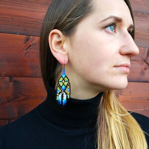 May include: A woman wearing a black turtleneck sweater with long, blonde hair. She is wearing a pair of long, beaded earrings with a geometric pattern in shades of blue, yellow, and black.
