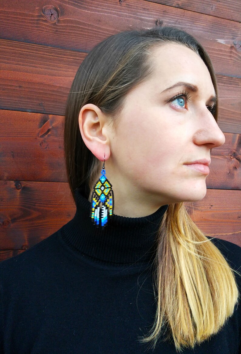 May include: A woman wearing a black turtleneck sweater with long, blonde hair. She is wearing a pair of long, beaded earrings with a geometric pattern in shades of blue, yellow, and black.