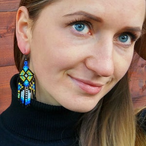 May include: A woman wearing a black turtleneck sweater with long brown hair. She is wearing a pair of long, beaded earrings with blue, yellow, and white accents.