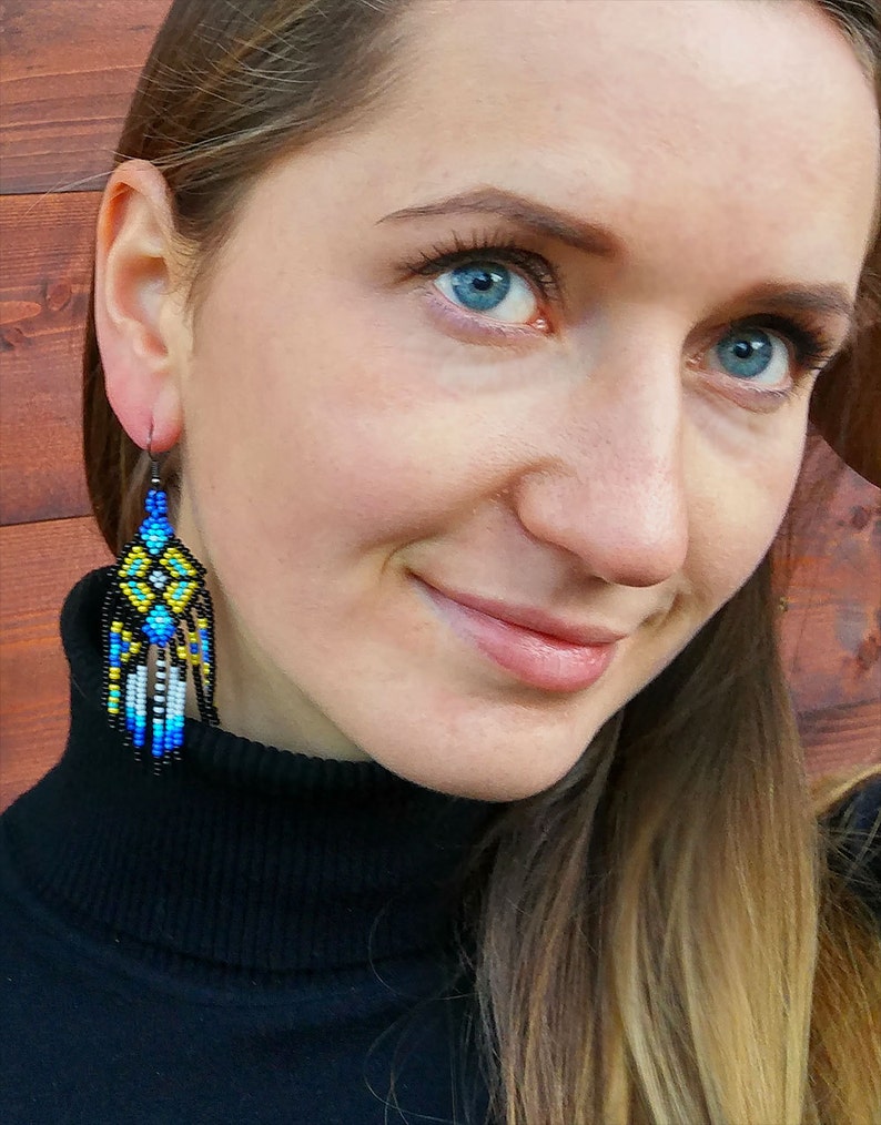 May include: A woman wearing a black turtleneck sweater with long brown hair. She is wearing a pair of long, beaded earrings with blue, yellow, and white accents.