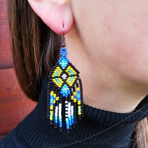 May include: A pair of dangle earrings with a geometric pattern of black, blue, white, and yellow beads. The earrings are made with a black metal hook.