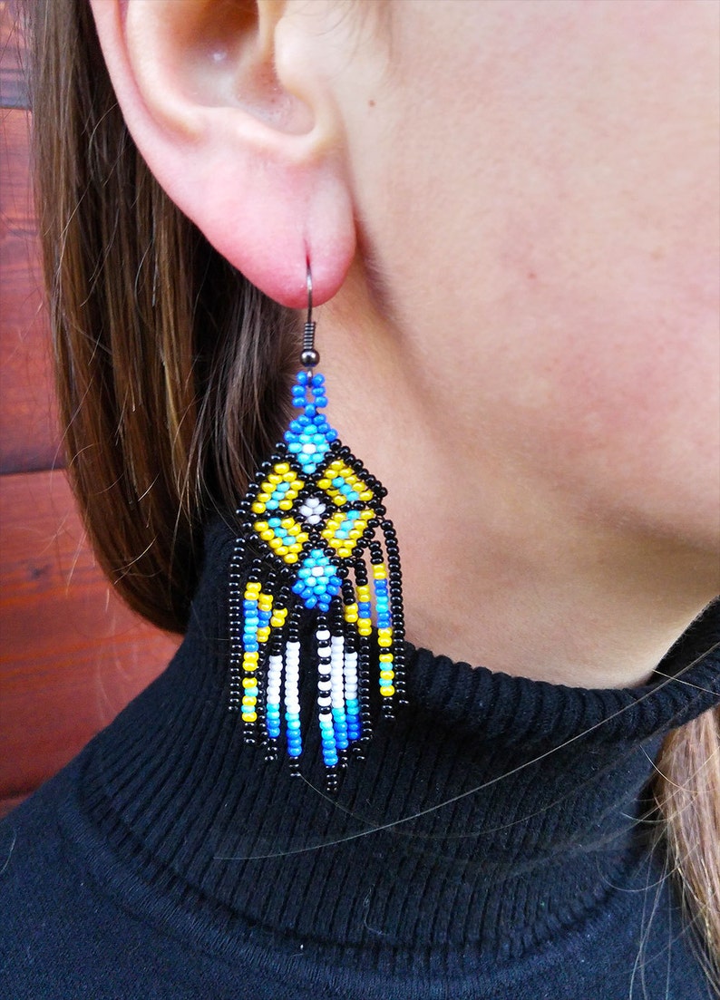 May include: A pair of dangle earrings with a geometric pattern of black, blue, white, and yellow beads. The earrings are made with a black metal hook.