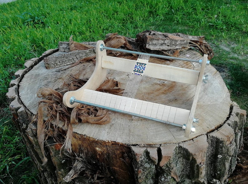 May include: A wooden loom with a beaded design in progress. The loom is made of natural wood and has a metal rod for weaving. The loom is sitting on a tree stump.