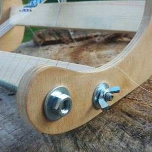 May include: A wooden loom with a metal rod and a white string attached to it. The loom has two metal nuts and bolts on the side.