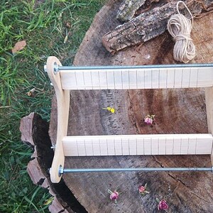 May include: A wooden loom with two sets of parallel bars, held together by metal rods. The loom is sitting on a tree stump with a ball of twine in the background.