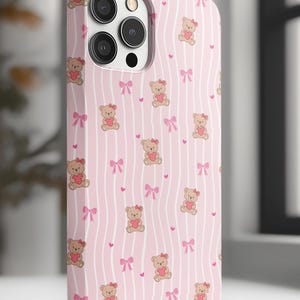 May include: Pink phone case with a repeating pattern of teddy bears holding hearts, pink bows, and small pink hearts on a striped background. The case has a camera cutout and is designed for a smartphone.