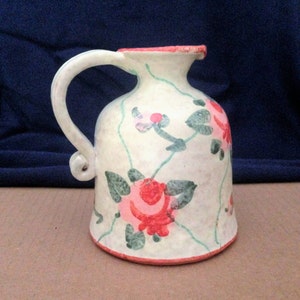 May include: A white ceramic pitcher with a curved handle and a flared rim. The pitcher is decorated with pink and red roses, green leaves, and green vines. The rim and base are trimmed in red.