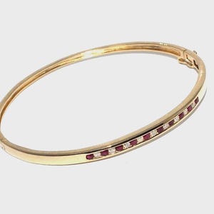 May include: A gold hinged bangle bracelet featuring a channel-set row of alternating round red rubies and clear white diamonds. The bracelet has a polished metal finish and a secure clasp mechanism visible on the right side of the band.