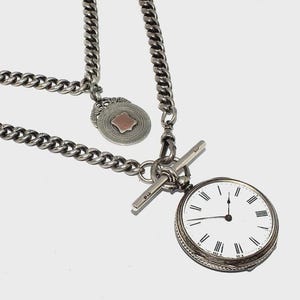 Puede incluir: Un reloj de bolsillo plateado con esfera blanca y números romanos negros unido a una gruesa cadena de eslabones. La cadena cuenta con un cierre de barra en T y un colgante circular con un detalle cuadrado central en metal envejecido.