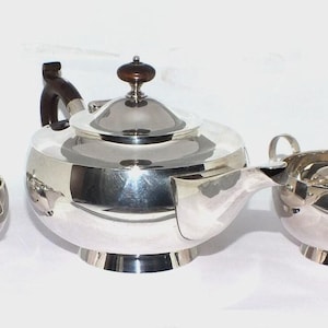 Peut inclure: Un service à thé en métal argenté composé de trois pièces : une théière avec une anse et un bouton de couvercle marron foncé, un sucrier et un pot à lait. Chaque pièce présente une finition métallique polie, un corps arrondi et une base.