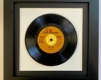 May include: Framed vintage 45 RPM vinyl record of "Foxy Lady" by The Jimi Hendrix Experience. The record is black with a yellow and orange label, set against a white mat and framed in a black square frame. The text "Reprise Records" is visible.
