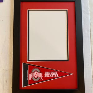 May include: A framed Ohio State Buckeyes pennant. The black frame surrounds a red mat with a white opening. The pennant is red with the Ohio State logo and the words "OHIO STATE BUCKEYES."