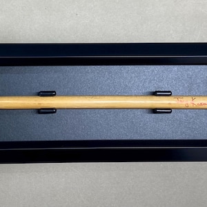 May include: A wooden drumstick signed by "T.I.  Akin" in red ink, displayed in a black frame with a dark blue mat.
