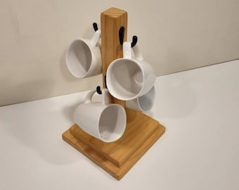 May include: A light-colored wooden mug tree with a square base and a central vertical post featuring four black metal hooks. Four plain white ceramic mugs are hanging from the hooks, each positioned to face outward from the central wooden stand.
