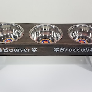 May include: A dark brown wooden elevated pet feeder stand featuring three circular stainless steel bowls. The front of the stand is personalized with the white text Bowser and Broccoli, each flanked by white paw print icons on both sides.