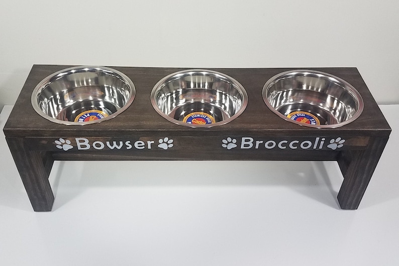 May include: A dark brown wooden elevated pet feeder stand featuring three circular stainless steel bowls. The front of the stand is personalized with the white text Bowser and Broccoli, each flanked by white paw print icons on both sides.