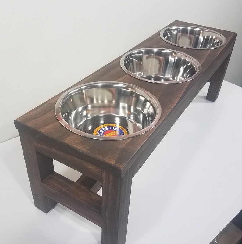 May include: A rectangular dark brown wooden stand featuring three circular cutouts, each holding a removable stainless steel bowl. The first bowl contains a circular sticker with the text Cadet Pet Bowls. The stand is supported by four legs.