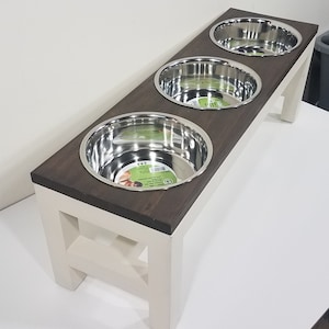 May include: A raised pet feeder stand featuring a dark brown rectangular wooden top with three circular cutouts holding removable stainless steel bowls. The base is constructed from off-white painted wood with four sturdy legs and cross-bracing.