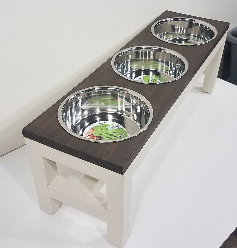 May include: A raised pet feeder stand featuring a dark brown rectangular wooden top with three circular cutouts holding removable stainless steel bowls. The base is constructed from off-white painted wood with four sturdy legs and cross-bracing.