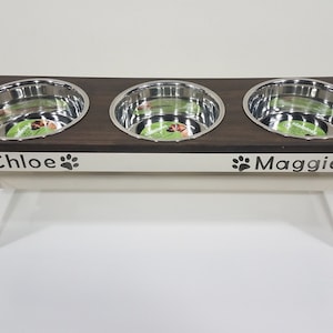 May include: A raised pet feeding station with a dark brown wooden top and cream-colored base. It features three circular stainless steel bowls and the names Chloe and Maggie printed on the front, each flanked by two black paw print icons.