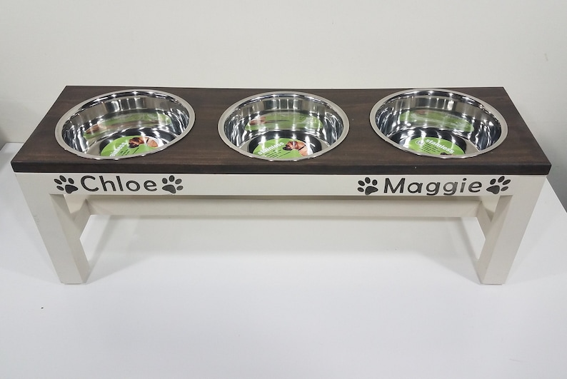 May include: A raised pet feeding station with a dark brown wooden top and cream-colored base. It features three circular stainless steel bowls and the names Chloe and Maggie printed on the front, each flanked by two black paw print icons.