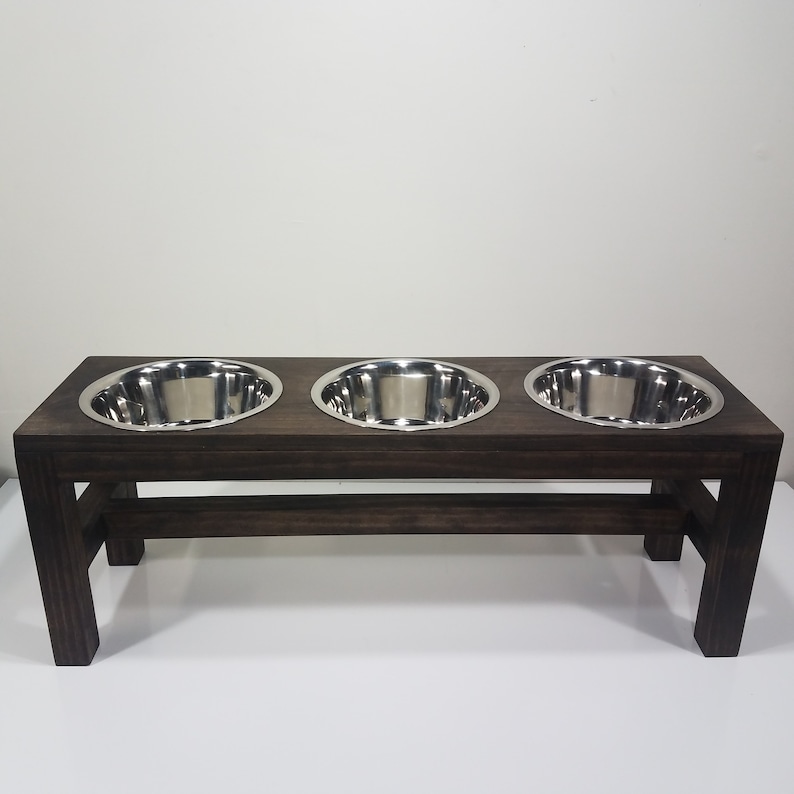 May include: A rectangular dark brown wooden stand featuring three circular cutouts, each holding a polished stainless steel bowl. The elevated feeder has four sturdy legs and a horizontal support bar connecting the front and back leg pairs.