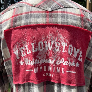 Puede incluir: Una camisa de franela roja con un patrón de cuadros blancos y grises. Un parche rojo está cosido en la parte posterior de la camisa con las palabras "Yellowstone National Park" en letras blancas. El texto "Wyoming Cody" también está impreso en blanco en el parche.