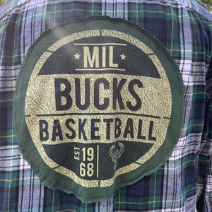 Puede incluir: Una camisa de franela a cuadros verde y blanca con un parche circular en la espalda. El parche es verde y presenta el texto "MIL BUCKS BASKETBALL EST 1968" en blanco.