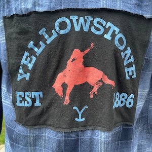 Puede incluir: Una camisa de franela a cuadros azules con un parche negro cosido en la espalda. El parche presenta una silueta roja de un vaquero montando un caballo, con el texto "Yellowstone Est 1886" en letras azules alrededor de la imagen. Una "Y" azul se encuentra debajo del caballo.