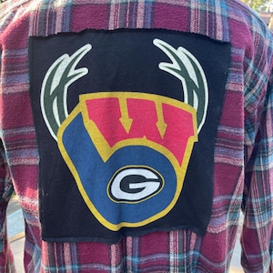 May include: A burgundy, blue, and green plaid flannel shirt with a black patch on the back. The patch features a logo with a blue, red, and yellow design, including a white "G" inside a blue circle.
