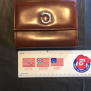 May include: A brown leather wallet with a silver C-shaped clasp. The wallet is closed and sitting on a black surface.