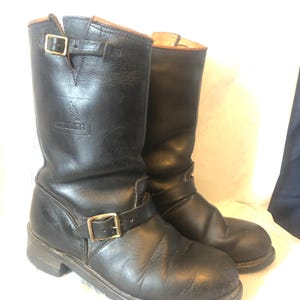 Puede incluir: Un par de botas de moto de cuero negro con cierres de hebilla. Las botas tienen una punta redonda y un tacón corto. La parte superior de las botas está forrada con cuero marrón. Las botas tienen un logotipo en el lateral.