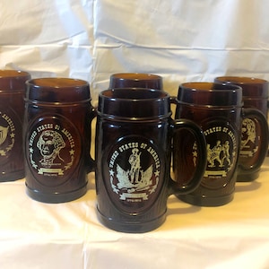 May include: Six brown glass mugs with handles. Each mug features a different image of an American historical figure or symbol, including George Washington, a soldier, and the American eagle. The mugs are decorated with the text "United States of America" and the years "1776-1976".