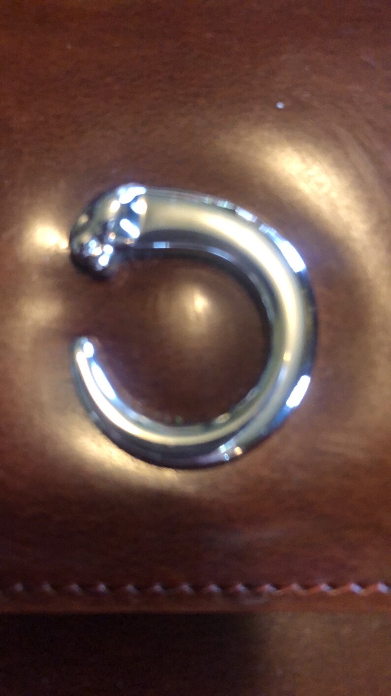 May include: A silver metal ring shaped like the letter C on a brown leather surface.