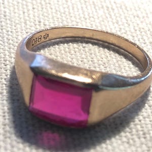 Peut inclure: Une bague dorée avec une pierre rectangulaire de couleur magenta. La bague a un design classique avec un anneau lisse et poli. La bague porte l'inscription "0 B" et un symbole de croix gravé sur l'anneau.