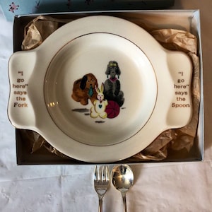 Può includere: Un piatto in ceramica bianca per bambini con un bordo dorato e un'immagine di un cane, un coniglio e un barboncino. Il piatto ha manici su entrambi i lati e il testo "I go here" says the Fork" sul manico sinistro e "I go here" says the Spoon" sul manico destro. Il piatto è in una scatola bianca con una fodera a motivi blu e rosa. Due forchette d'argento sono su una superficie bianca.