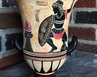 May include: A ceramic vase with two handles, painted in a black and white silhouette style with a red and orange accent. The vase depicts a Greek warrior holding a shield and a spear. The vase has a brown base and a cream colored body.
