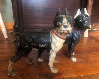以下が含まれることがあります： 黒と白の鋳鉄製のボストン・テリアのドアストッパー2個。1匹の犬は頭を横に傾けて立っており、もう1匹の犬は頭を前に向けて座っています。両方の犬は赤い首輪をしています。