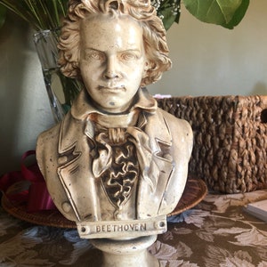 Può includere: Un busto in gesso bianco di Beethoven con un viso scolpito dettagliato e un colletto arricciato. Il busto è su un piedistallo bianco con la scritta "BEETHOVEN" incisa su una placca.