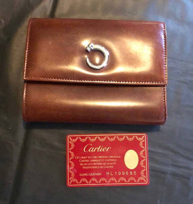 May include: A brown leather Cartier wallet with a silver C logo. The wallet is closed and sitting on a black surface. A red card with white text reads 'Cartier' and 'CET OBJET EST UNE CREATION ORIGINALE CARTIER, FABRIQUE ET CONTROLE SELON LES CRITERES DE QUALITE TRADITIONNELS DE CARTIER CUIRS/LEATHER HL 109695'.