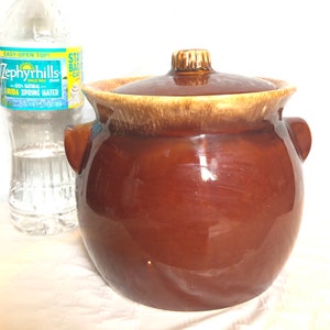 May include: A brown ceramic crock pot with a lid and two handles. The crock pot has a glazed finish and a light brown rim.