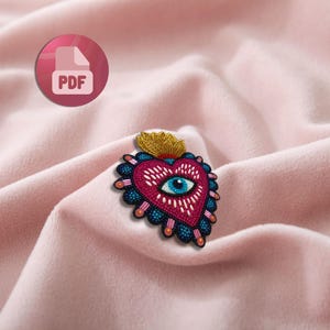 Op de afbeelding: Een hartvormige patch met een oogmotief. Het rode hart heeft witte stiksels, een blauw oog en een gouden kroon. Het is omgeven door een blauwe en oranje kralenrand, tegen een roze stoffen achtergrond.