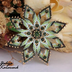 以下が含まれることがあります： ビーズで作られた花のブローチ。詳細なデザインです。ブローチの中央には濃い緑色の宝石があり、緑、白、金色の花びらに囲まれています。ブローチの直径は約7.6cmです。アーティストの名前、Yahya Karaenkは左下にあります。
