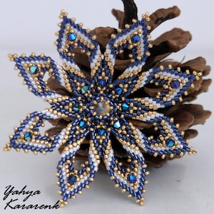 May include: A handcrafted, beaded flower ornament with a snowflake design. The ornament features navy blue, white, and gold beads, with iridescent blue accents. The ornament is resting on a pine cone. The artist's name, Yahya Kararenk, is in the lower left corner.