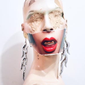 May include: A mannequin head displays long, silver-toned flame-shaped earrings. The mannequin features a collage element across the lower face, depicting a mouth with bright red lipstick and slightly parted lips, contrasting with the plain beige head.