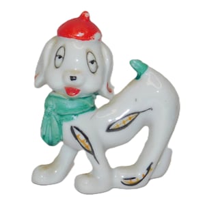 May include: A ceramic dog figurine with a red hat and a green scarf. The white dog has black spots and a red mouth. The figurine is a vintage collectible.