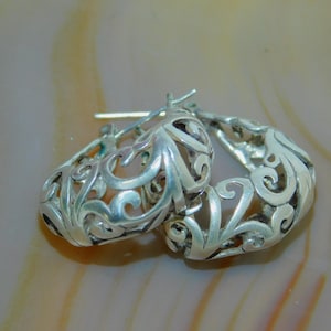 May include: A pair of silver hoop earrings featuring an intricate, openwork filigree design with swirling patterns. The earrings have a hinged post closure mechanism and a polished metallic finish that reflects light across the curved surfaces.