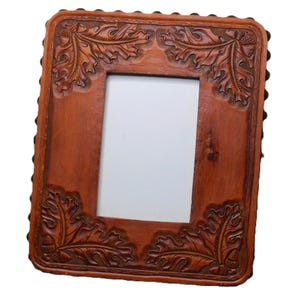 May include: A rectangular, brown leather picture frame with carved oak leaf and acorn designs in each corner. The frame's edges are lined with small, dark studs. The center opening is white.